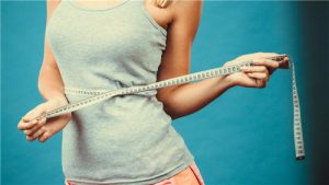 Fit fitness girl measuring her waistline with measure tape on blue.