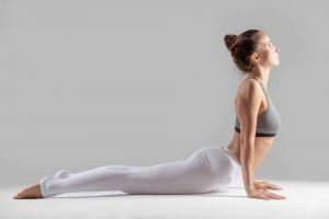 Young attractive woman practicing yoga, stretching in Cobra exercise, Bhujangasana pose, working out wearing sportswear, indoor full length, isolated against grey studio background.