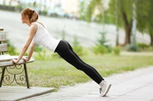 Young woman performing push-ups from a bench in a park.