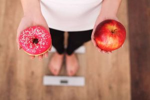 Woman Measuring Body Weight On Weighing Scale Holding Donut and apple.