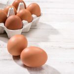 Close-up view of raw chicken eggs in egg box on white wooden background.