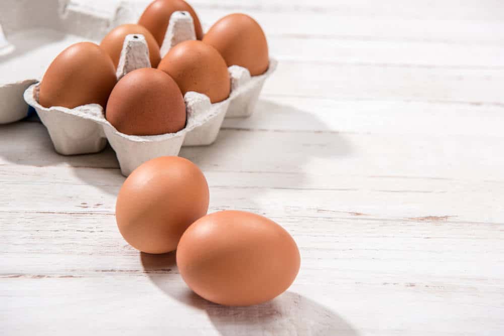 Close-up view of raw chicken eggs in egg box on white wooden background.