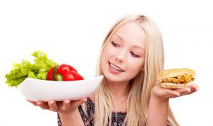 Thoughtful young woman holding a hamburger with chicken and plate with vegetables, isolated against white background.