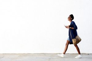 Full length side view portrait of trendy young black woman walking outdoors and listening to music on her mobile phone.